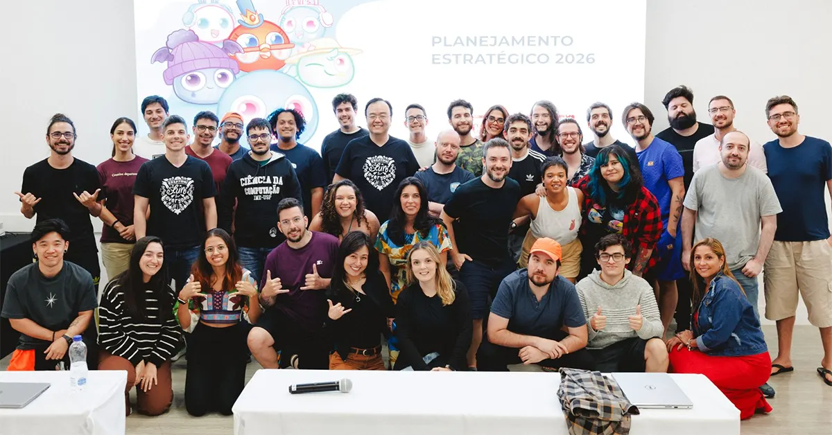 Foto de equipe reunida para planejamento estratégico, com grupo de pessoas em frente a um banner ao fundo escrito "Planejamento Estratégico 2026" e ilustração de robôs e personagens. Mesa com itens de escritório na frente.