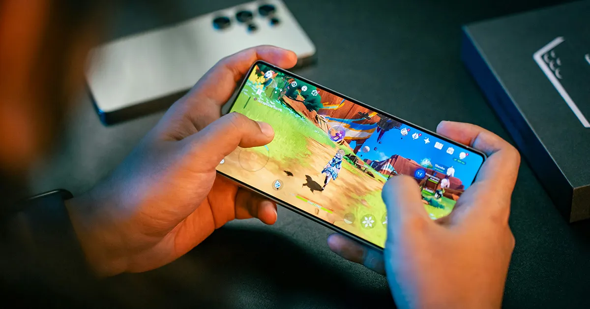 Pessoa usando um smartphone para jogar em ambiente de escritório ou estúdio, com a tela mostrando um jogo colorido e ícones na parte superior e inferior.